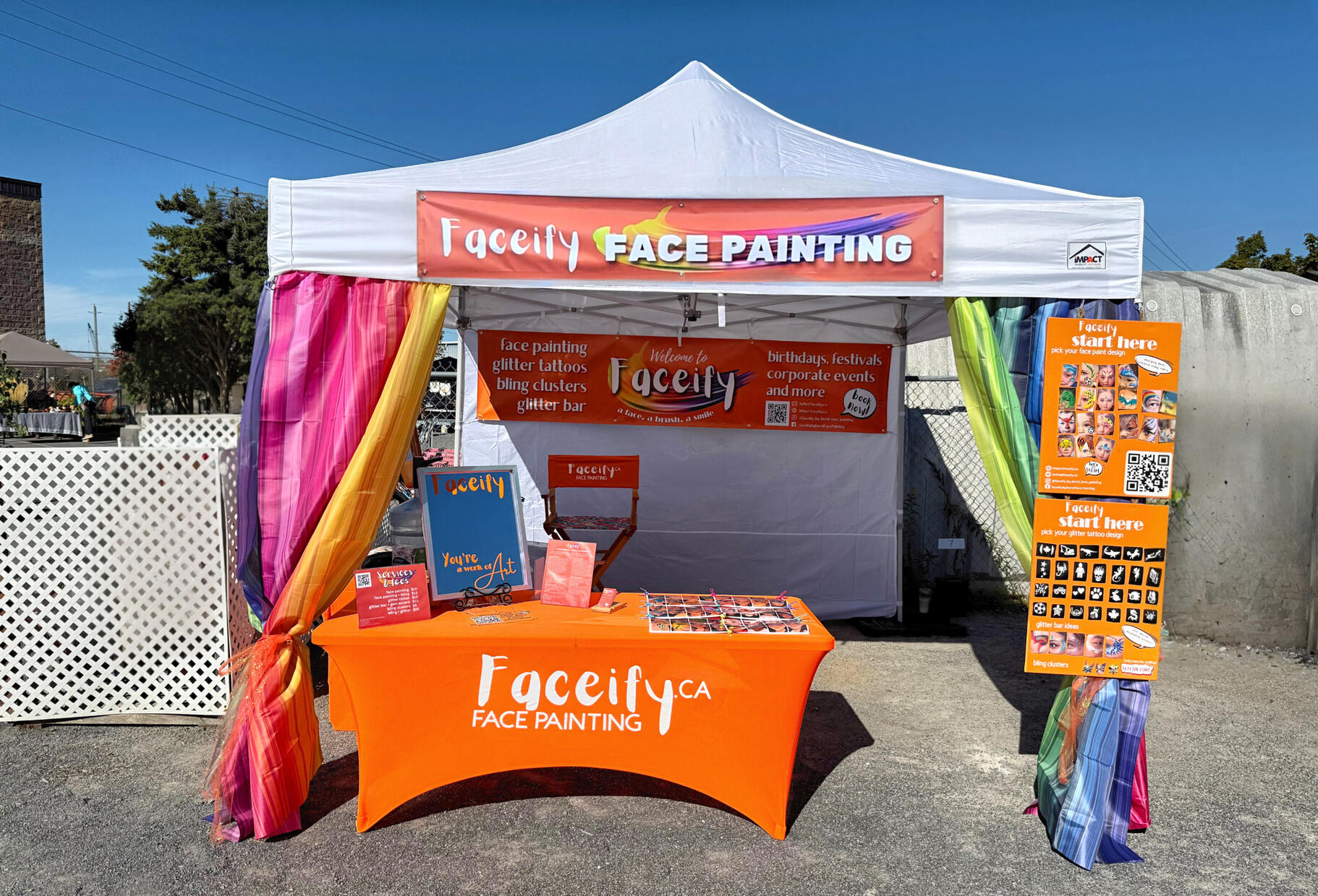 Faceify Ottawa face painting setup at an outdoor event, with design boards, glitter tattoo options, rainbow drapes and service table